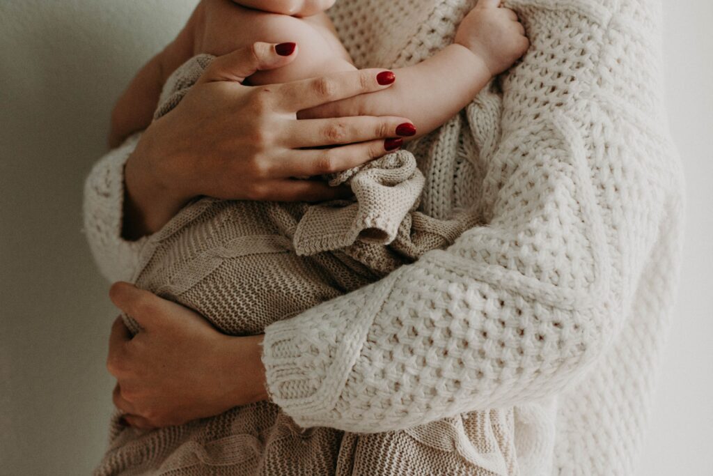 Une mère vêtue d'un pull beige, tient son bébé dans les bras. Ses ongles sont peints en rouge. 